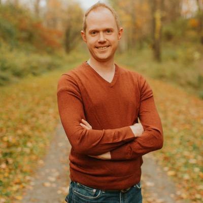 Image includes a picture of Aric Bryant standing in the middle of a road with a fall foliage background. He has his arms crossed too. He is also wearing an orange sweater and a pair of blue jeans.