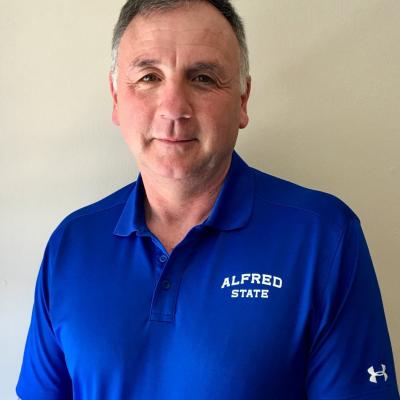 Joseph Bailey wearing an Alfred State polo shirt, standing against a neutral background, smiling warmly at the camera.