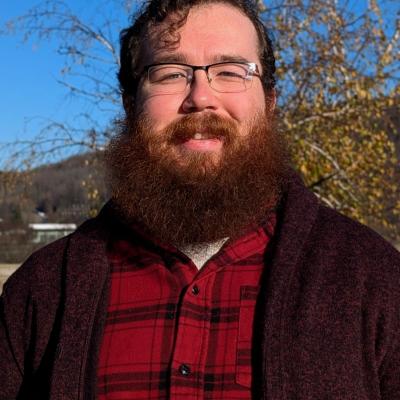 Jeremy Howard, a white man with curly brown hair and a mid-size beard, wearing glasses, a red flannel with black stripes, and a dark red cardigan, smiling in front of a tree, hill, and sky.
