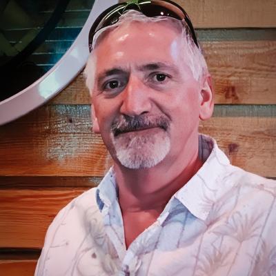 A casual portrait of a man with short gray hair and a mustache, wearing a patterned button-down shirt and sunglasses resting on his head. He is positioned in front of a wooden wall with part of a circular decorative object visible.