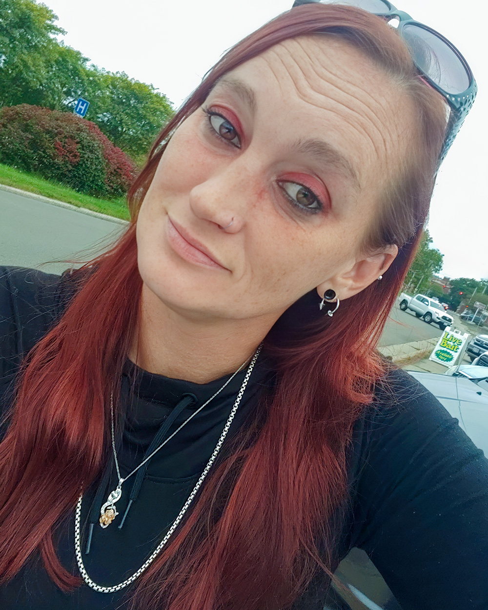 A casual outdoor portrait of A casual outdoor portrait of a woman with long red hair, wearing a black top, layered silver necklaces, and earrings. She has sunglasses resting on her head and is looking at the camera with a soft smile, set against a background of greenery and parked cars. with long red hair, wearing a black top, layered silver necklaces, and earrings. She has sunglasses resting on her head and is looking at the camera with a soft smile, set against a background of greenery and parked cars.