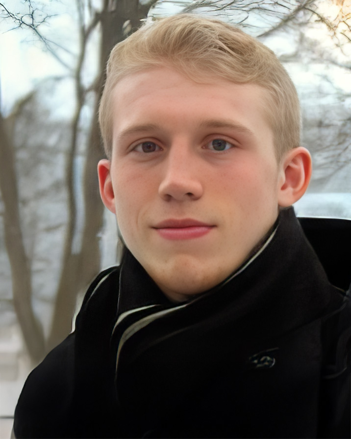 A professional portrait Blake Ilges, wearing a black scarf and coat. The background features an outdoor winter scene with bare trees and a soft, cool-toned atmosphere.