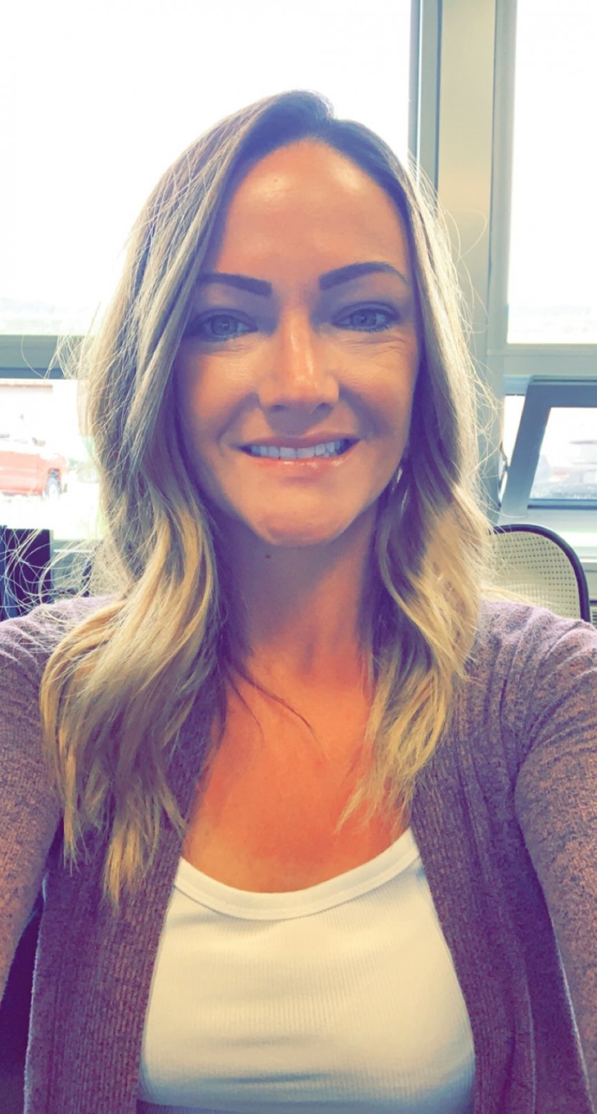 A photo of Rachel Brandow with blonde wavy hair, wearing a white top and lavender cardigan, sitting near a bright window with a soft natural light illuminating the scene.