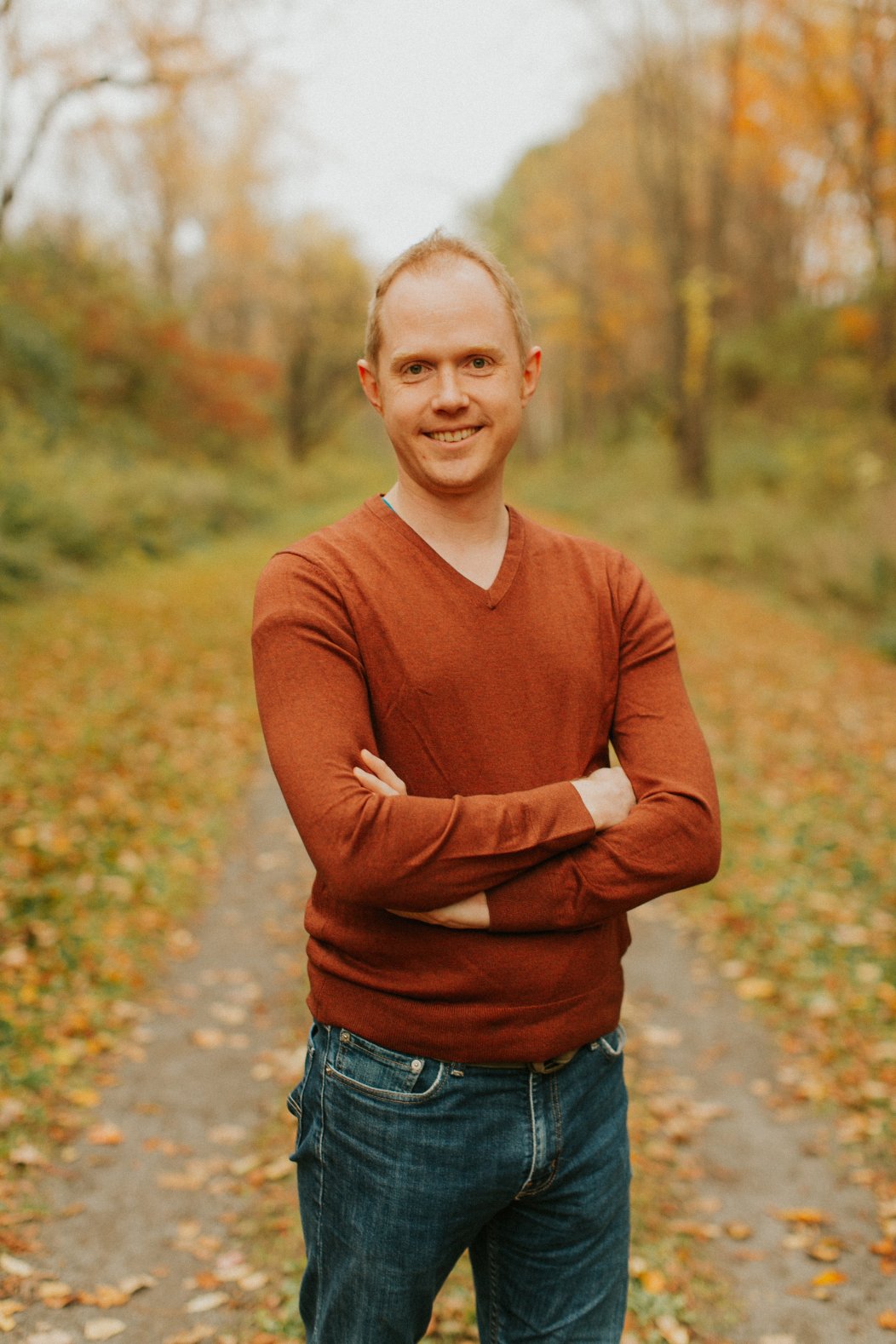 Image includes a picture of Aric Bryant standing in the middle of a road with a fall foliage background. He has his arms crossed too. He is also wearing an orange sweater and a pair of blue jeans.
