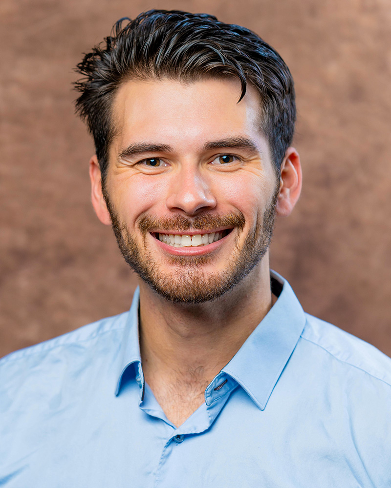 A professional headshot Chance Olivas with neatly styled dark hair and a trimmed beard, wearing a light blue collared shirt. The background is a warm, textured brown, providing a neutral and polished setting for the portrait.