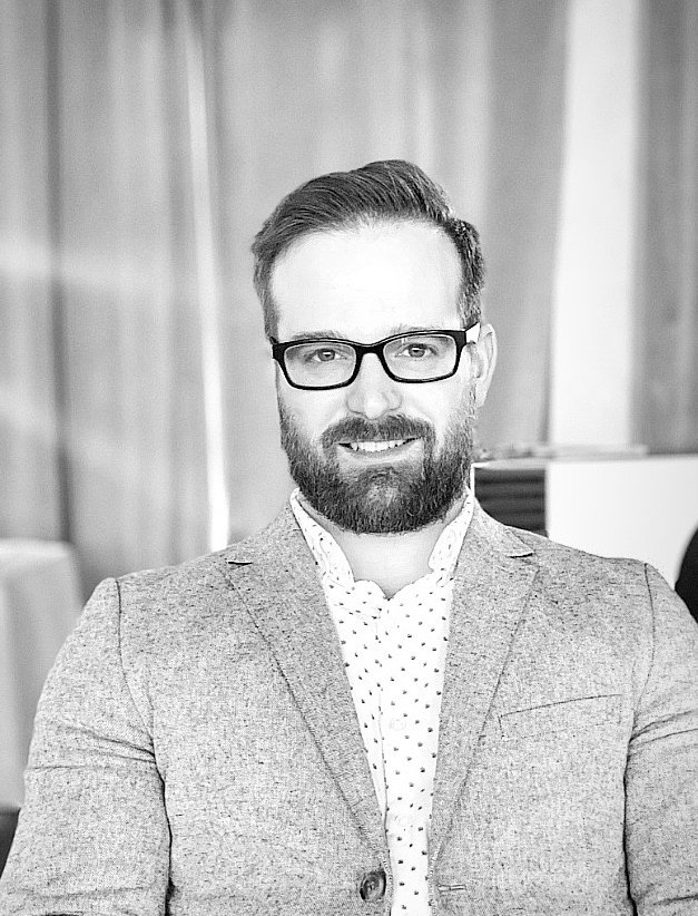 A grayscale photo of Michael Chapman wearing glasses, a patterned shirt, and a light blazer, seated in a well-lit indoor setting with soft curtains in the background.