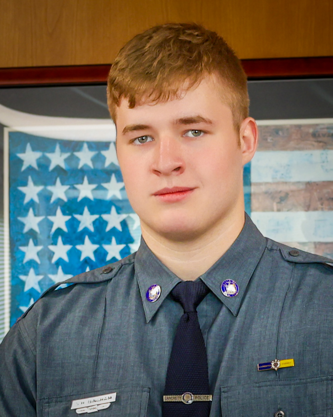 A university police officer poses in uniform in front of a stylized American flag backdrop.