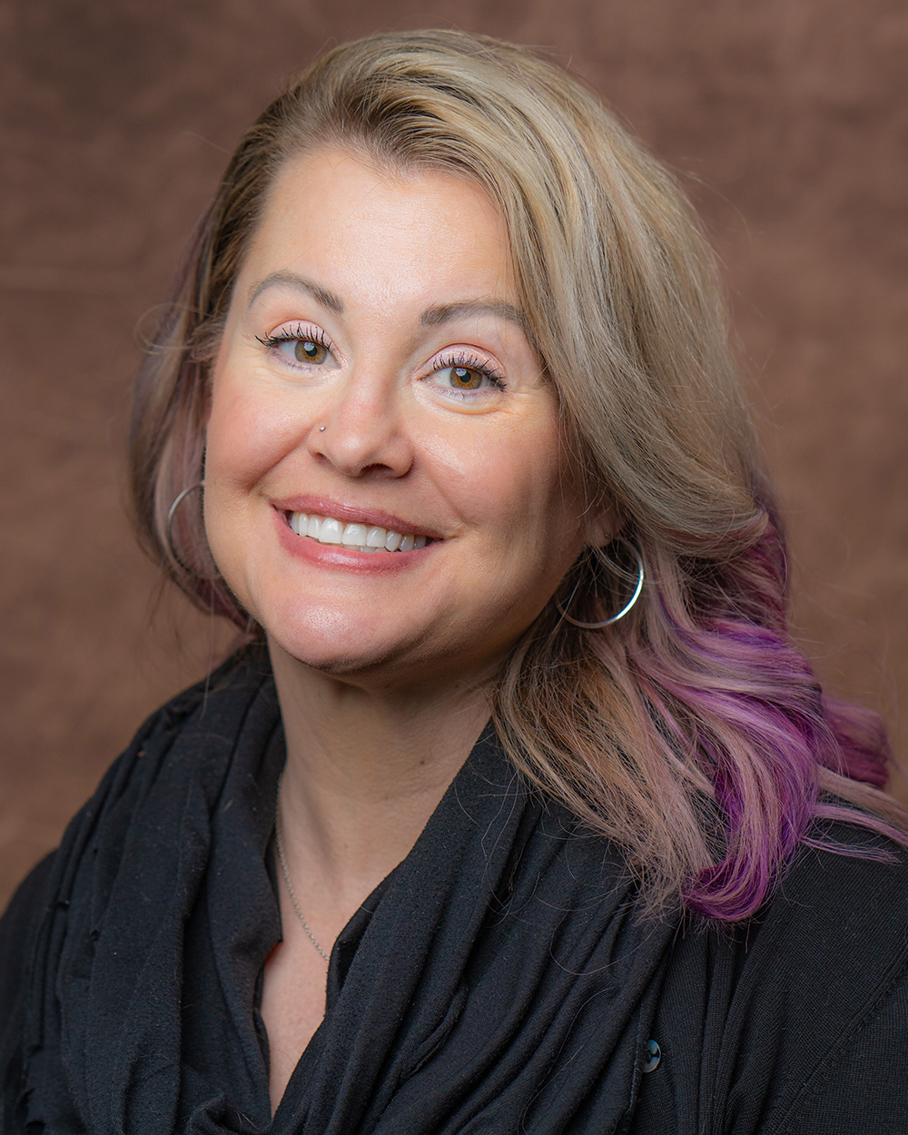 A professional portrait of Del Ray Honeycutt with blonde and purple hair, wearing hoop earrings and a black scarf. The background is a textured brown, giving the image a warm and formal feel.