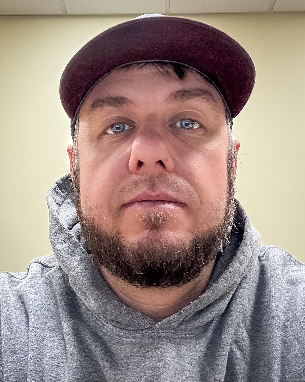A portrait of William Deming III with a trimmed beard, wearing a maroon baseball cap and a gray hoodie. He gazes directly at the camera against a plain, light background.