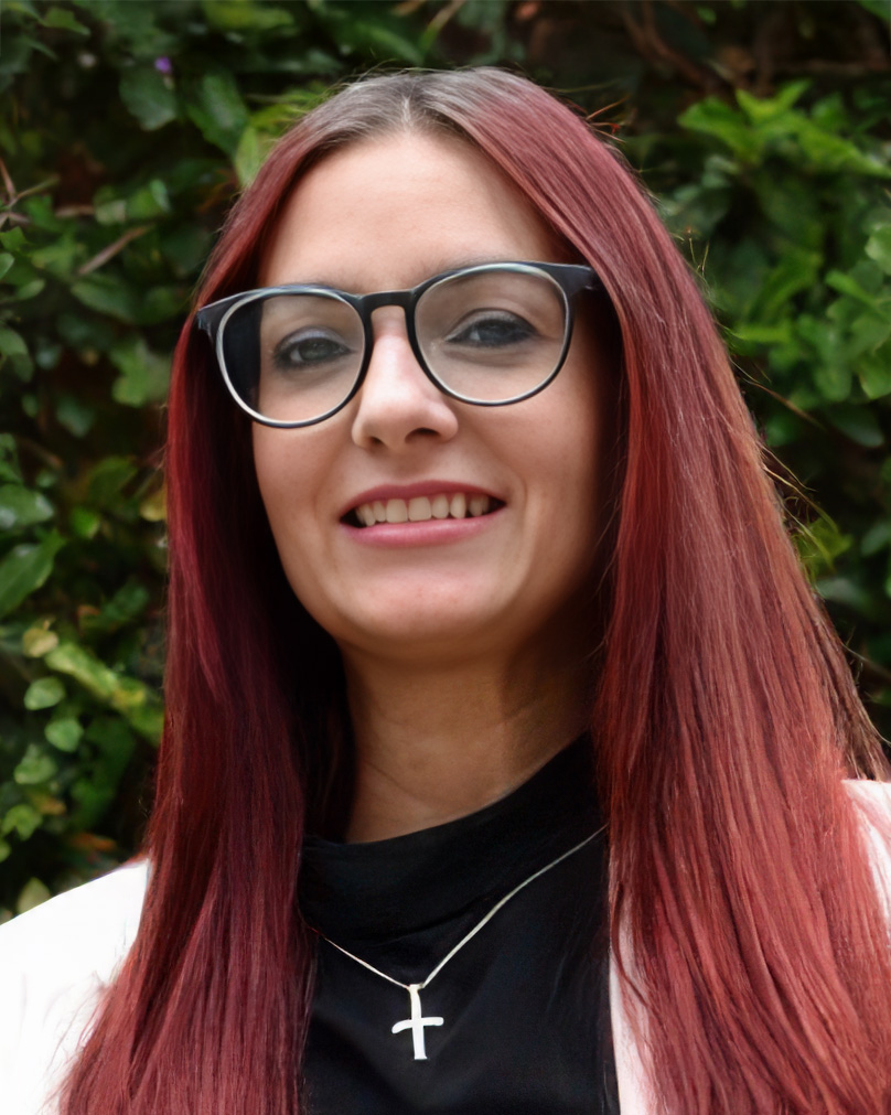 A portrait of Tiffany Demiris with straight, shoulder-length red hair wearing glasses. She is smiling and dressed in a black top paired with a white blazer. A silver cross necklace adorns her neck.
