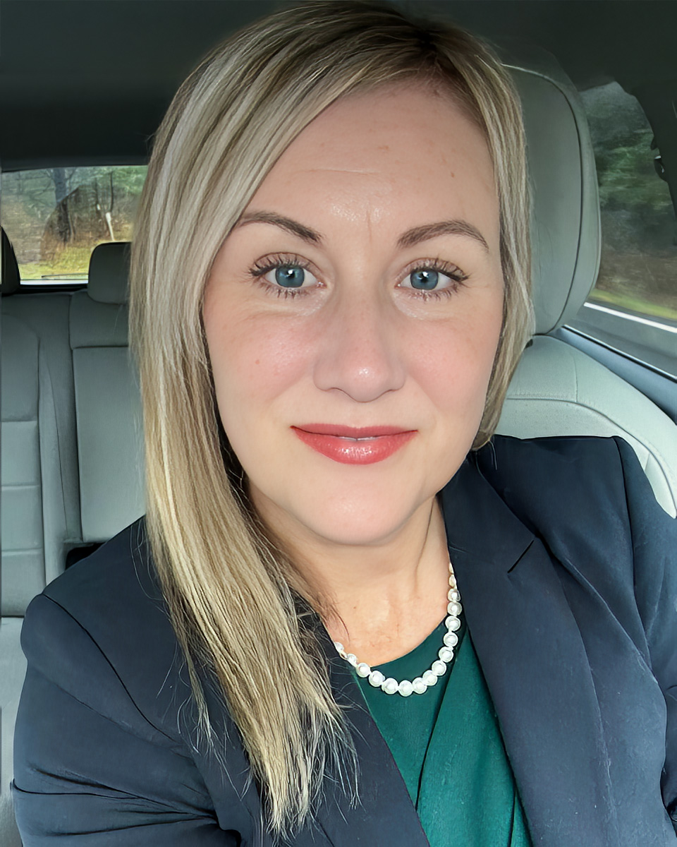 A professional portrait of a woman seated in a car, wearing a dark blazer, a green top, and a pearl necklace. She has light blonde hair and a friendly smile, with a soft outdoor background visible through the car windows.