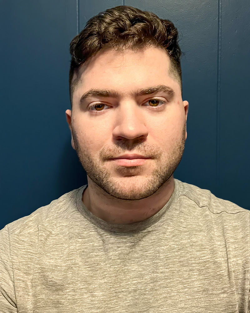 A headshot of Eric Scouten with short, wavy brown hair and a neatly groomed beard. He is wearing a light gray shirt and is set against a solid, dark blue background, creating a clean and modern aesthetic.