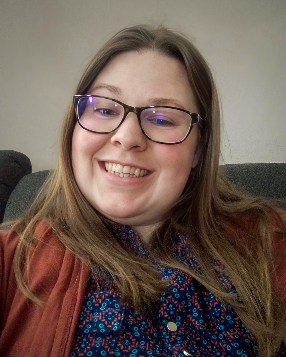 A smiling woman wearing glasses and a patterned top with a rust-colored cardigan takes a photo indoors. The neutral background and her cheerful expression create a warm and approachable vibe.