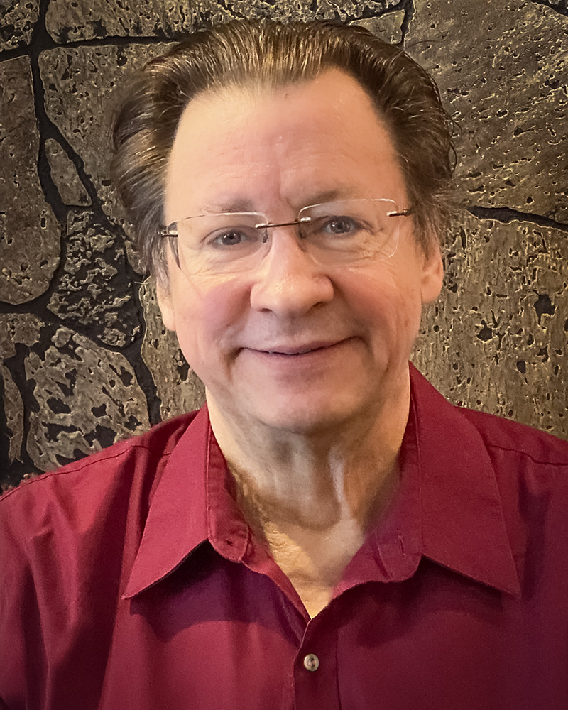 A headsot of Leroy Hanchett with neatly combed hair and glasses, wearing a burgundy button-up shirt.