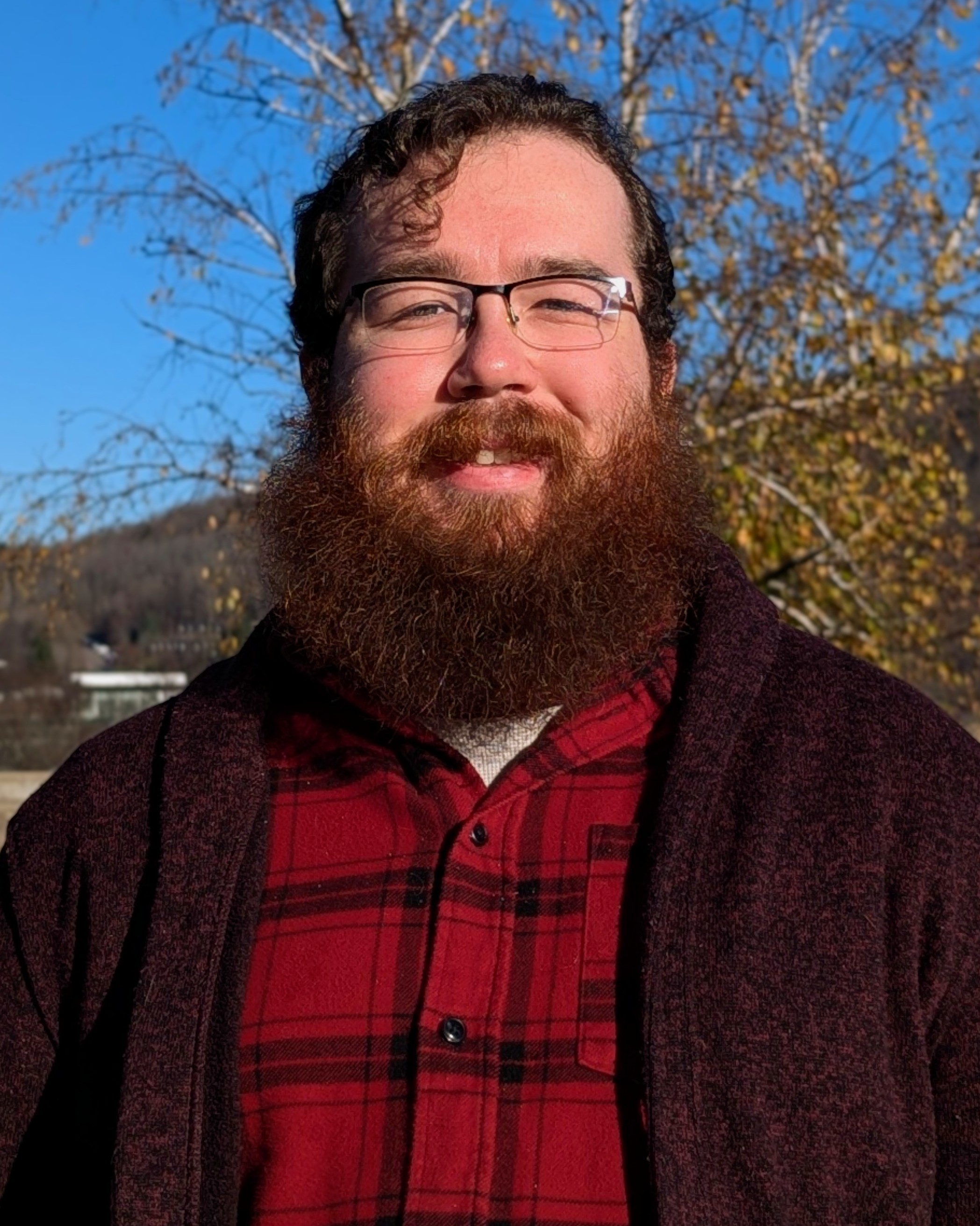 Jeremy Howard, a white man with curly brown hair and a mid-size beard, wearing glasses, a red flannel with black stripes, and a dark red cardigan, smiling in front of a tree, hill, and sky.
