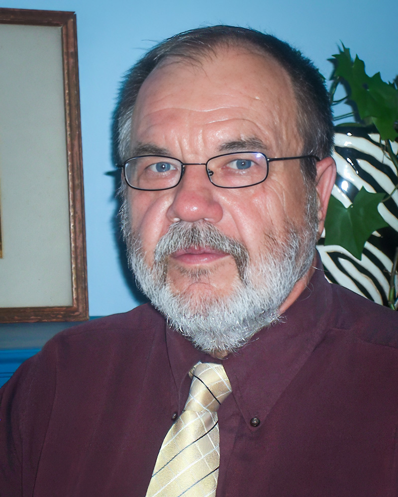 Headshot of Steven Jakobi with glasses and a white beard, wearing a maroon shirt and yellow tie. The background includes a blue wall and greenery.