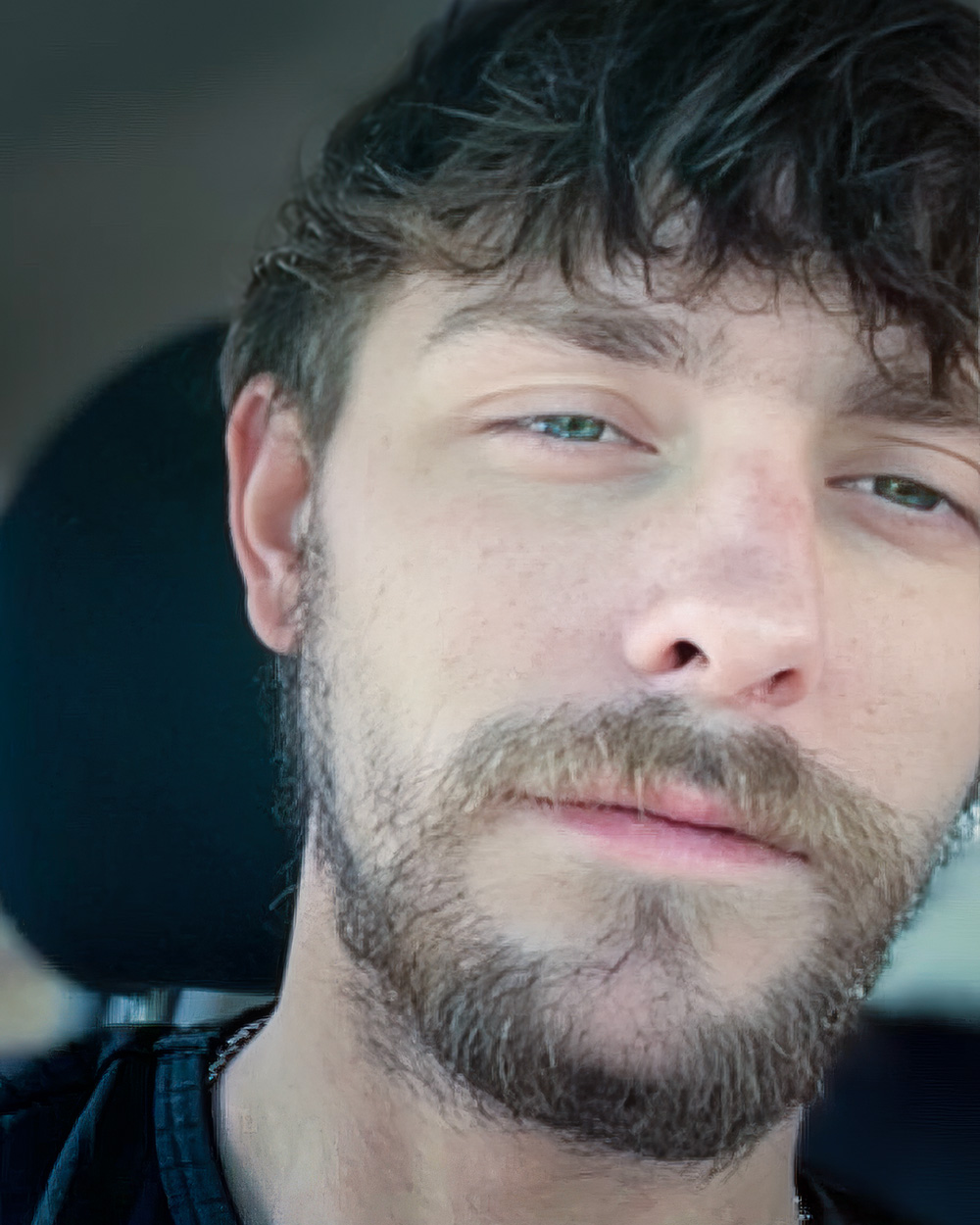 A close-up portrait of Kenneth Moore with a beard and tousled hair. His expression is relaxed, and he is photographed against the interior of a car, softly lit.