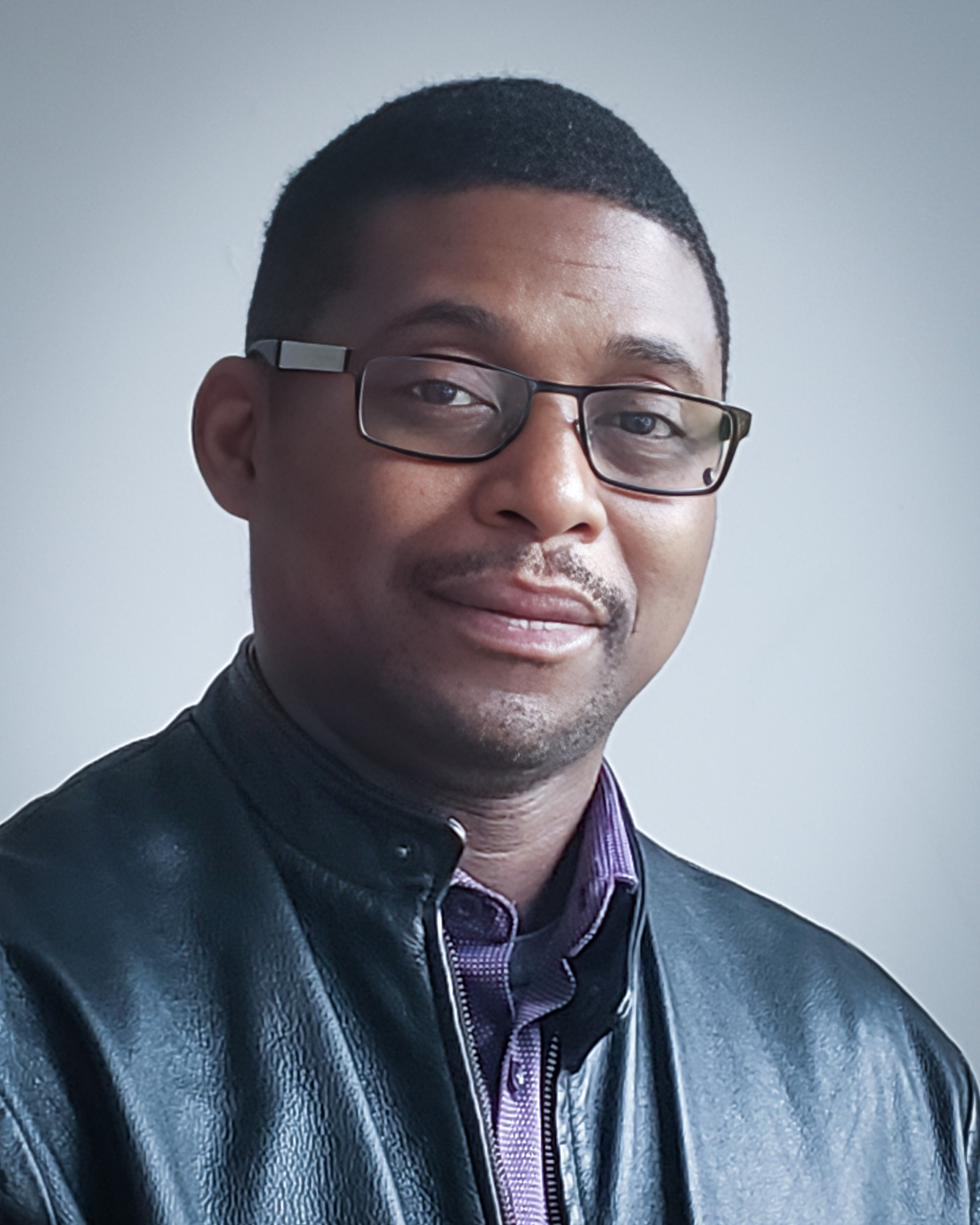A professional portrait of James Oyedapo wearing glasses, a black leather jacket, and a purple collared shirt on a neutral background.