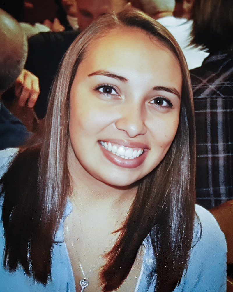 A photo of Cassandra Parker, wearing a light blue shirt and a heart-shaped necklace. The background features a group of people in a casual setting.