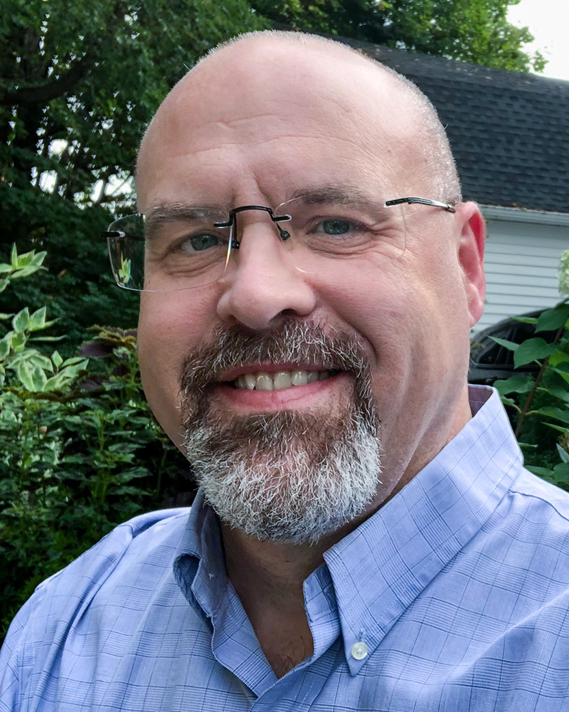 Kevin Payne with glasses and a goatee, smiling warmly. He is dressed in a light blue shirt, with a backyard featuring greenery and hydrangeas in the background.