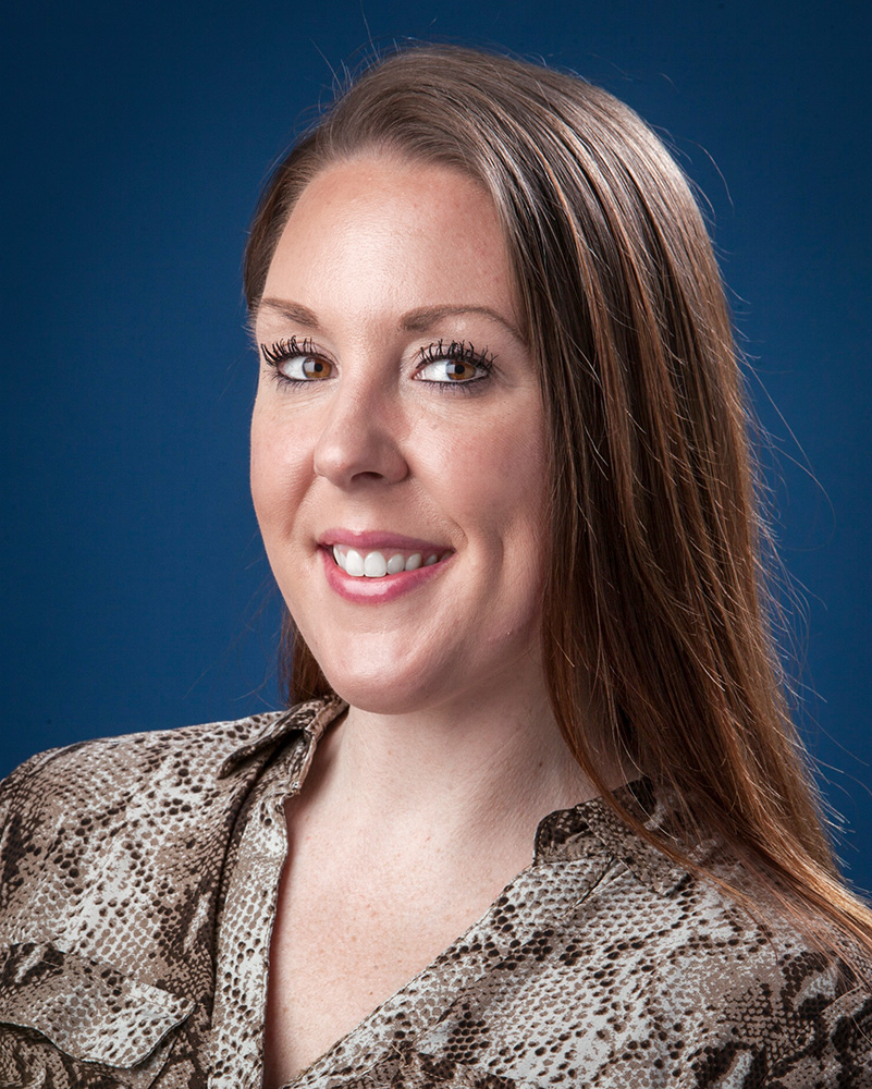 A photo of Christine Reynolds with long brown hair, wearing a patterned blouse. The background is a solid blue.