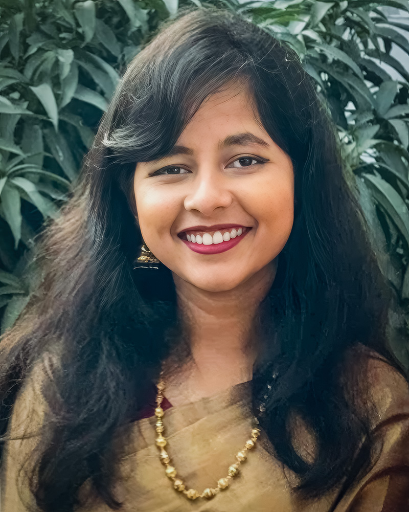 A portrait of Saki Rezwana with long, dark hair, adorned with gold earrings and a necklace. She is dressed in an elegant beige saree and smiles warmly, with a lush green backdrop enhancing the lively atmosphere.