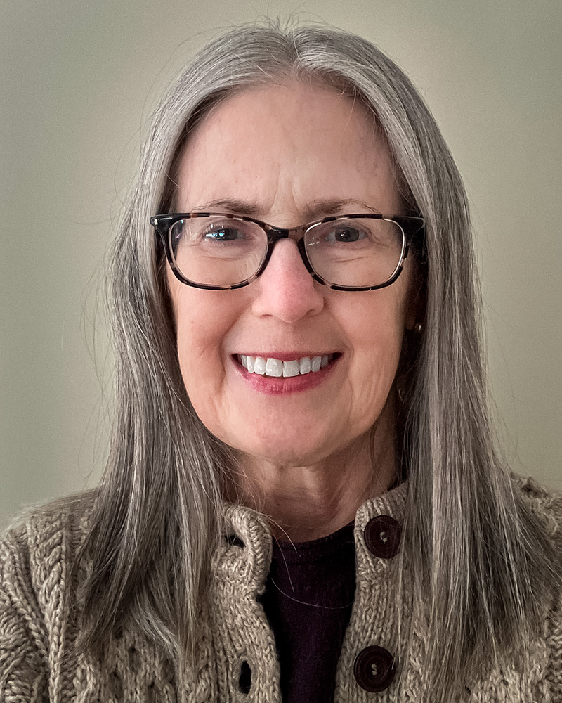 A portrait of Sandra Rigas with straight gray hair and glasses. She is wearing a cozy knitted sweater with large buttons, standing against a neutral background.