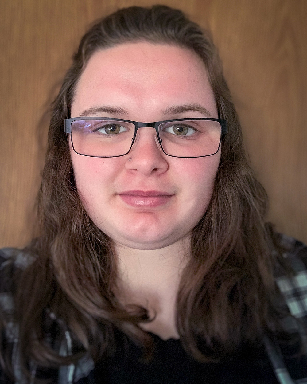A friendly portrait of Caitlin Stewart wearing glasses and a plaid shirt, set against a wooden background, creating a natural and welcoming vibe.