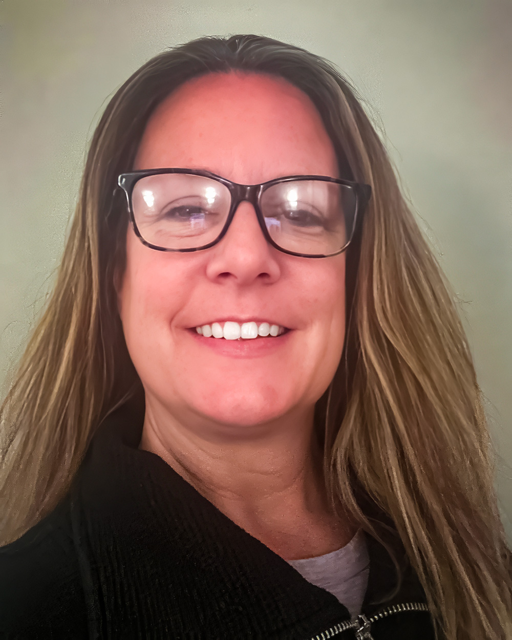 A portrait of Shyla Streb with straight, light brown hair, wearing glasses and a dark zip-up sweater. She smiles brightly against a plain, soft-toned background.