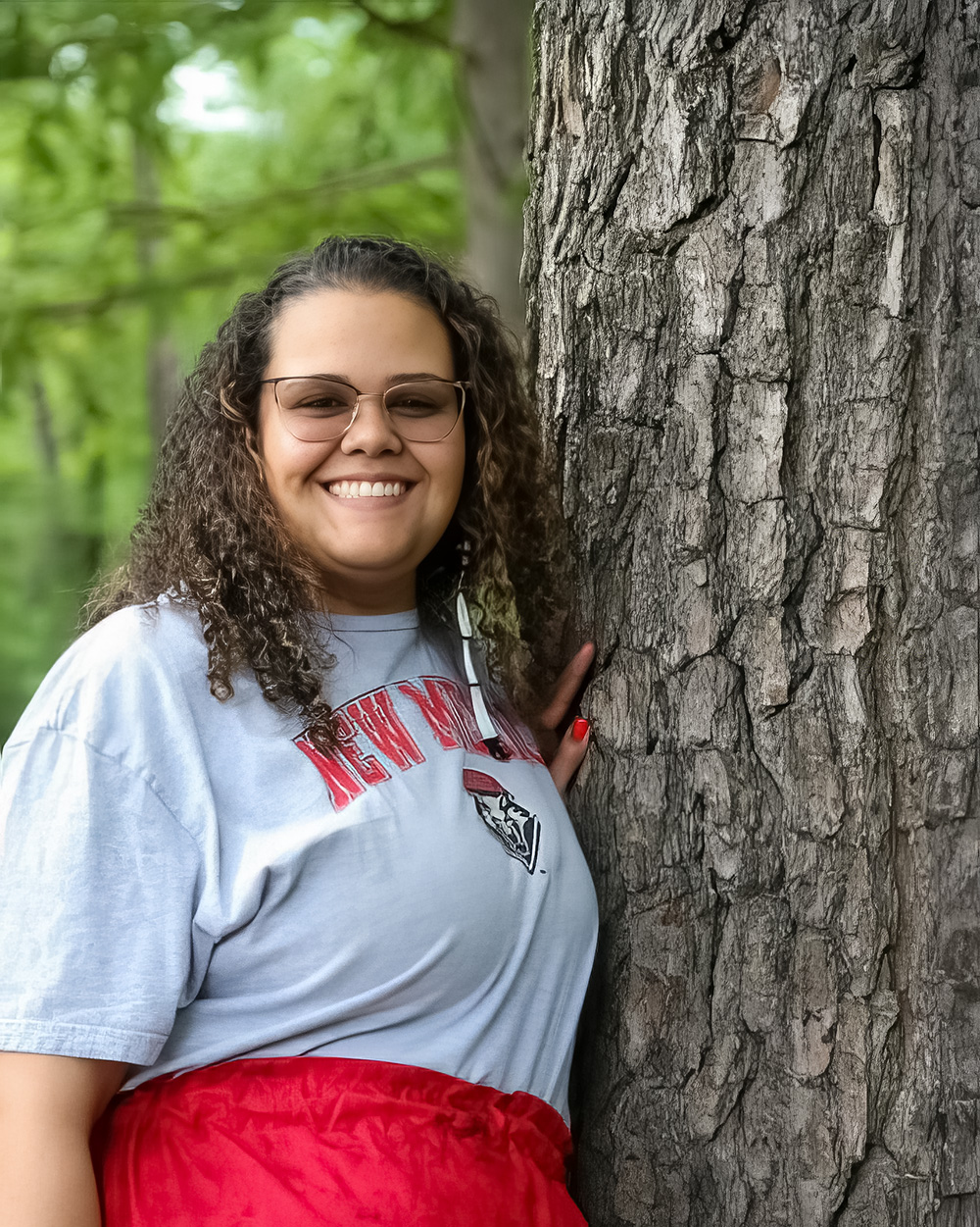 A photo of Taylor Strickland with curly hair smiles warmly, standing next to a large tree. She wears glasses, a light gray “New Mexico” T-shirt, and a bright red skirt. The lush green forest background adds to the serene, natural setting.