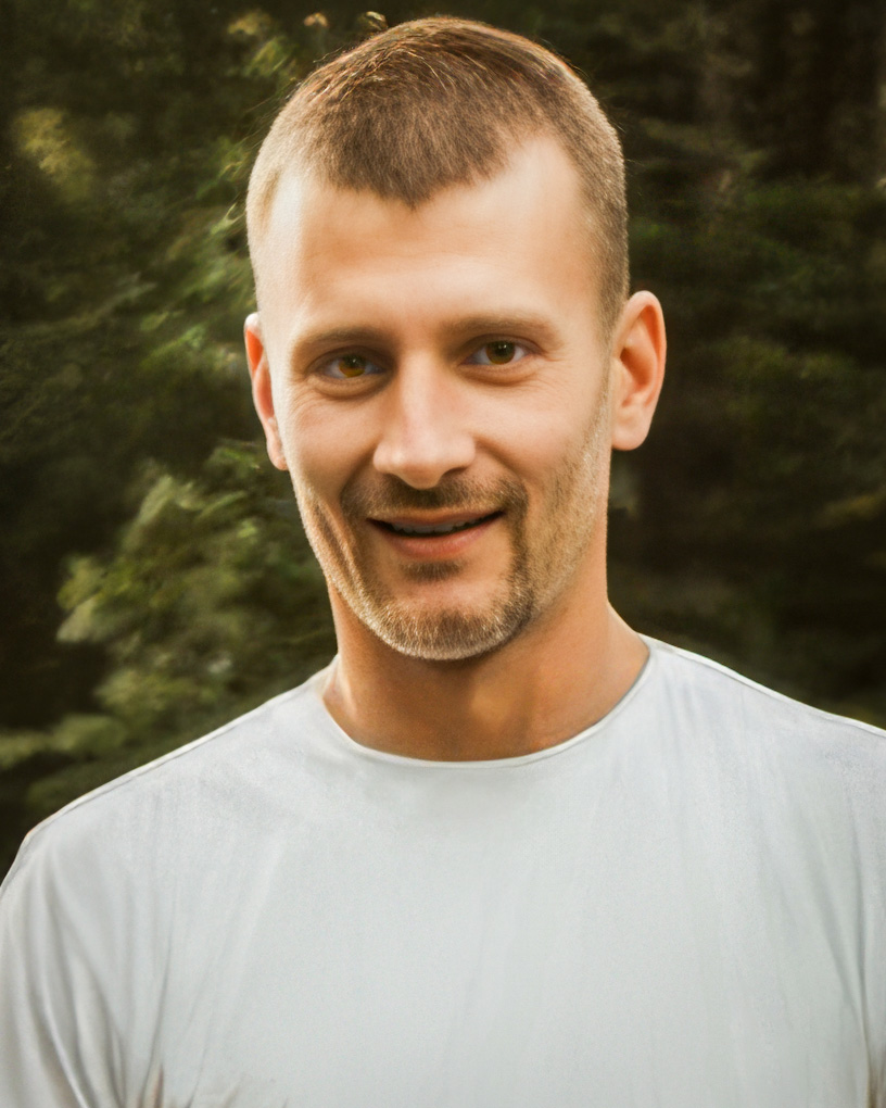 A photo of Kyle Wiedemann with short, light brown hair and a neatly trimmed beard smiles outdoors. He wears a white t-shirt, with a lush green forest in the softly blurred background.