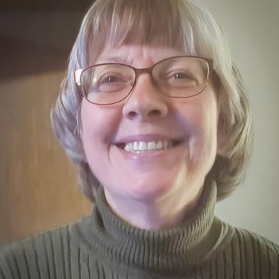 A headshot of Marilyn Robin with short, light gray hair and glasses, smiling warmly at the camera. They are wearing a ribbed olive-green turtleneck sweater, and the background features soft, neutral tones.