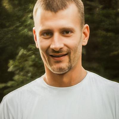 A photo of Kyle Wiedemann with short, light brown hair and a neatly trimmed beard smiles outdoors. He wears a white t-shirt, with a lush green forest in the softly blurred background.